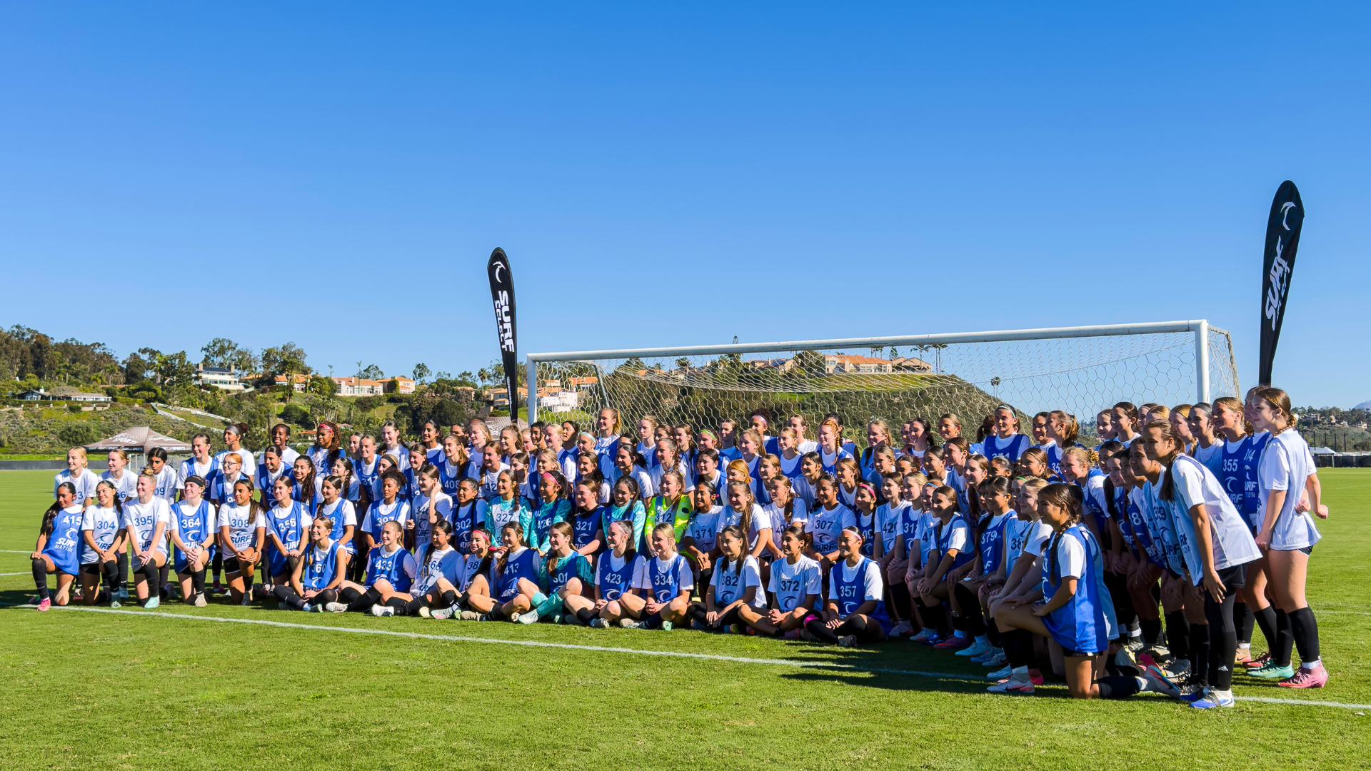 Large group of girls in blue jerseys posing for a team photo on a soccer field with the goal behind them.