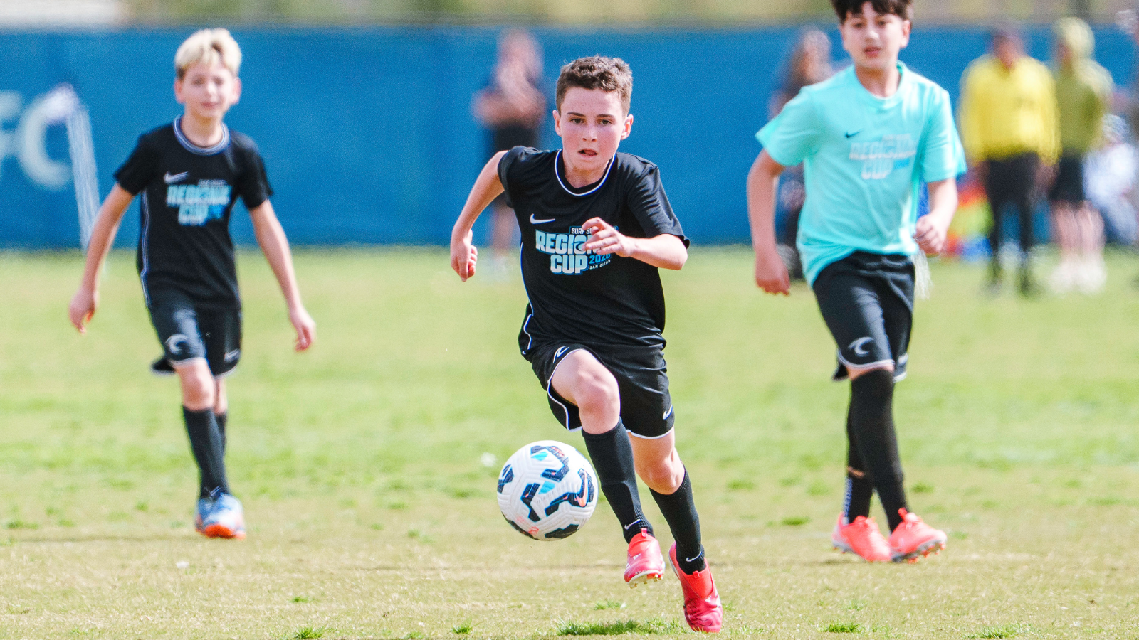 Three young boys play a soccer match on a sunny field; central child in black jersey chases a ball with two teammates nearby.