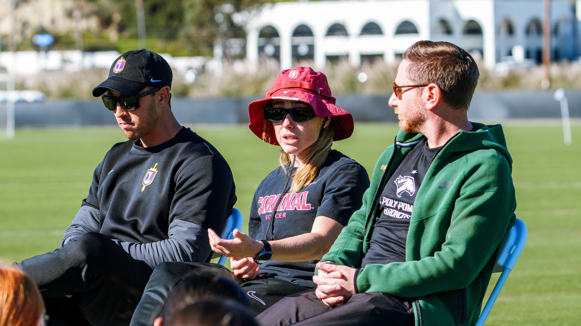 Three adults in athletic clothing sit on blue chairs on a sunny soccer field, wearing sunglasses and hats while talking.