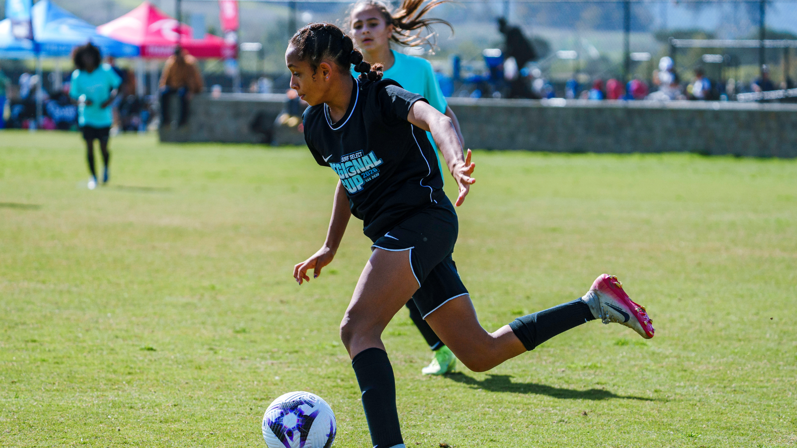 Girl in black soccer uniform chases a ball on a sunny field during a youth match, foreground action with teammates in background