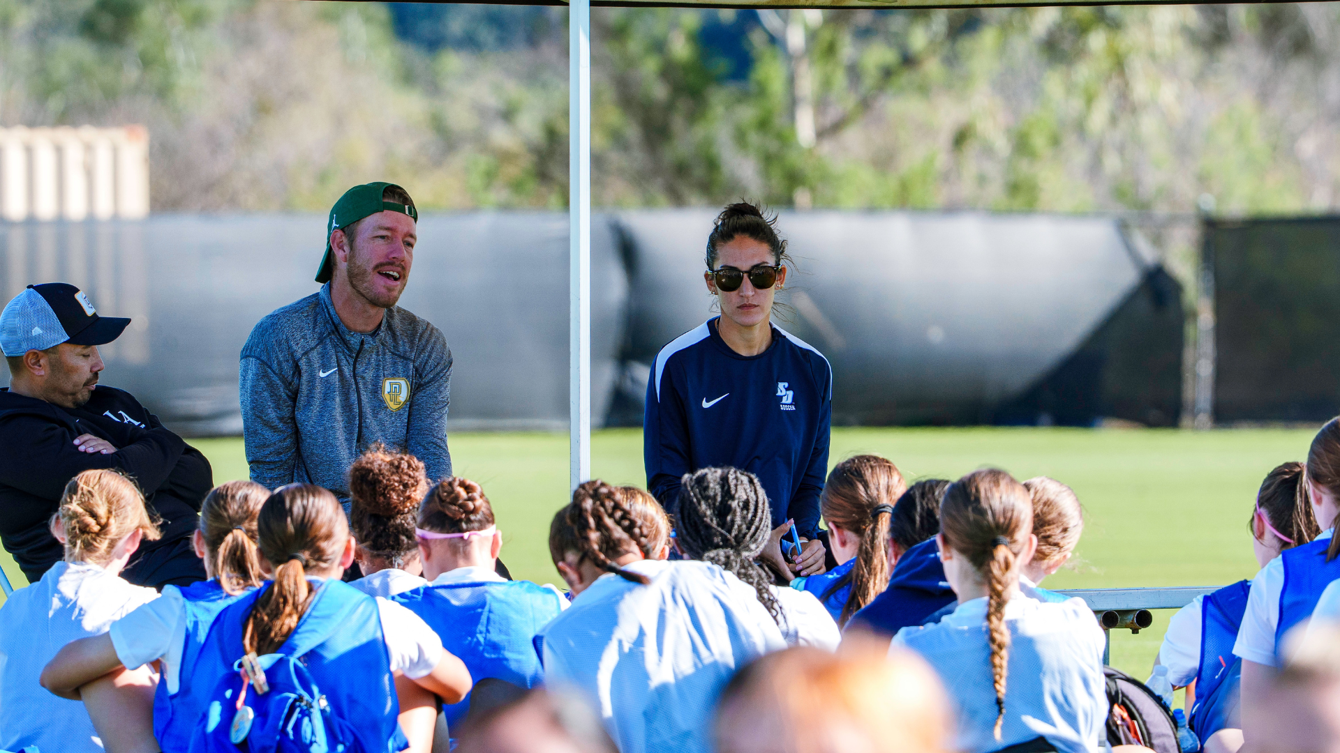Coach and assistant address a group of young players in blue pinnies under a canopy on a sunny sports field.