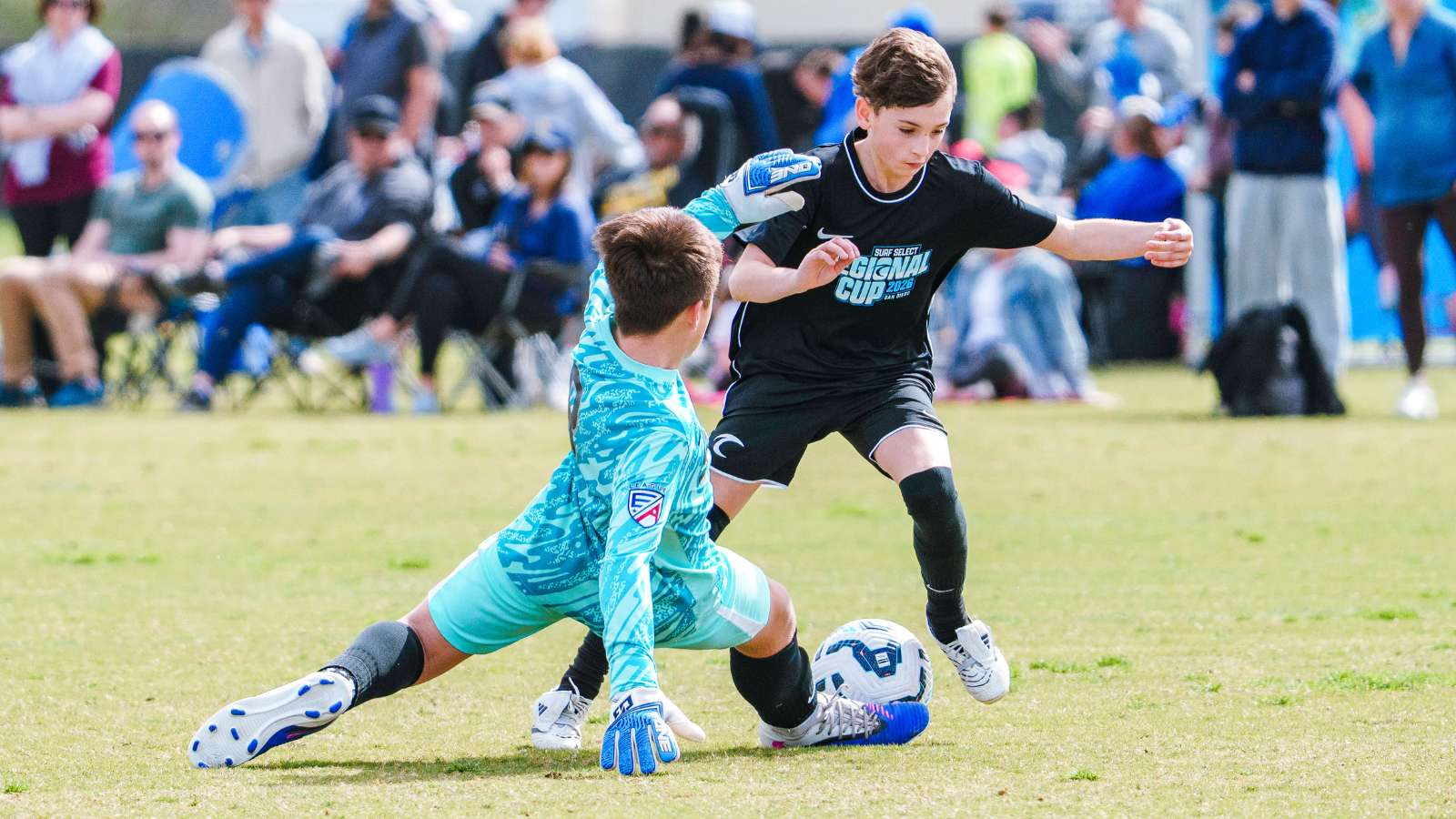 Two youth soccer players compete for the ball on a sunny field while a goalie in turquoise dives to block the shot, spectators watching in the background.