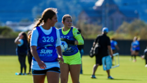 Two female field hockey players in blue pinnies on a sunny grass field, one holding a white ball and looking ahead.