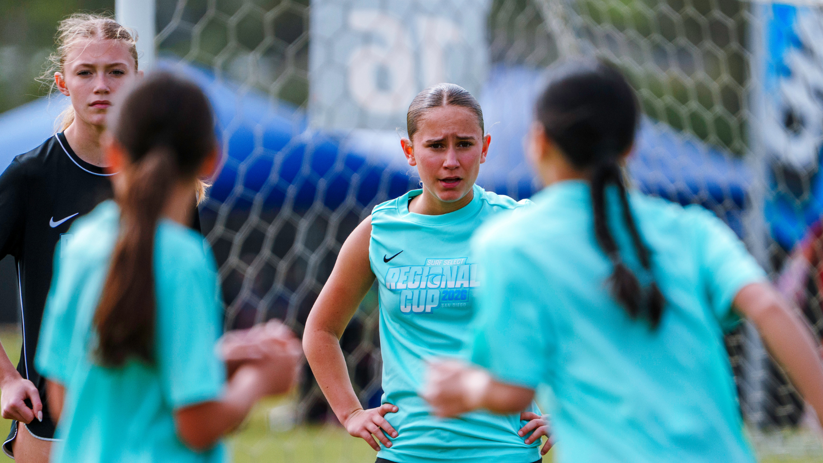 Teenage girls' soccer team in teal jerseys on a practice field, central player with hands on hips near the goal net.