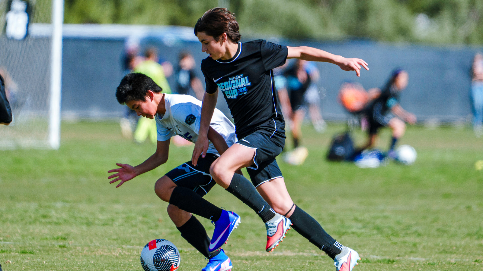 Two young soccer players chase the ball on a grassy field, one in black kit ahead of a white-team opponent near the ball.