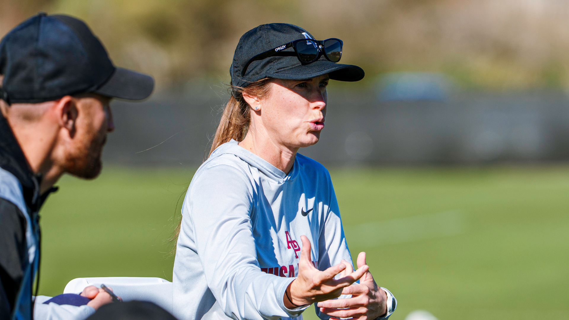 Woman in a light blue athletic shirt and cap speaks with animated hand gestures outdoors, a man sits nearby wearing a cap.