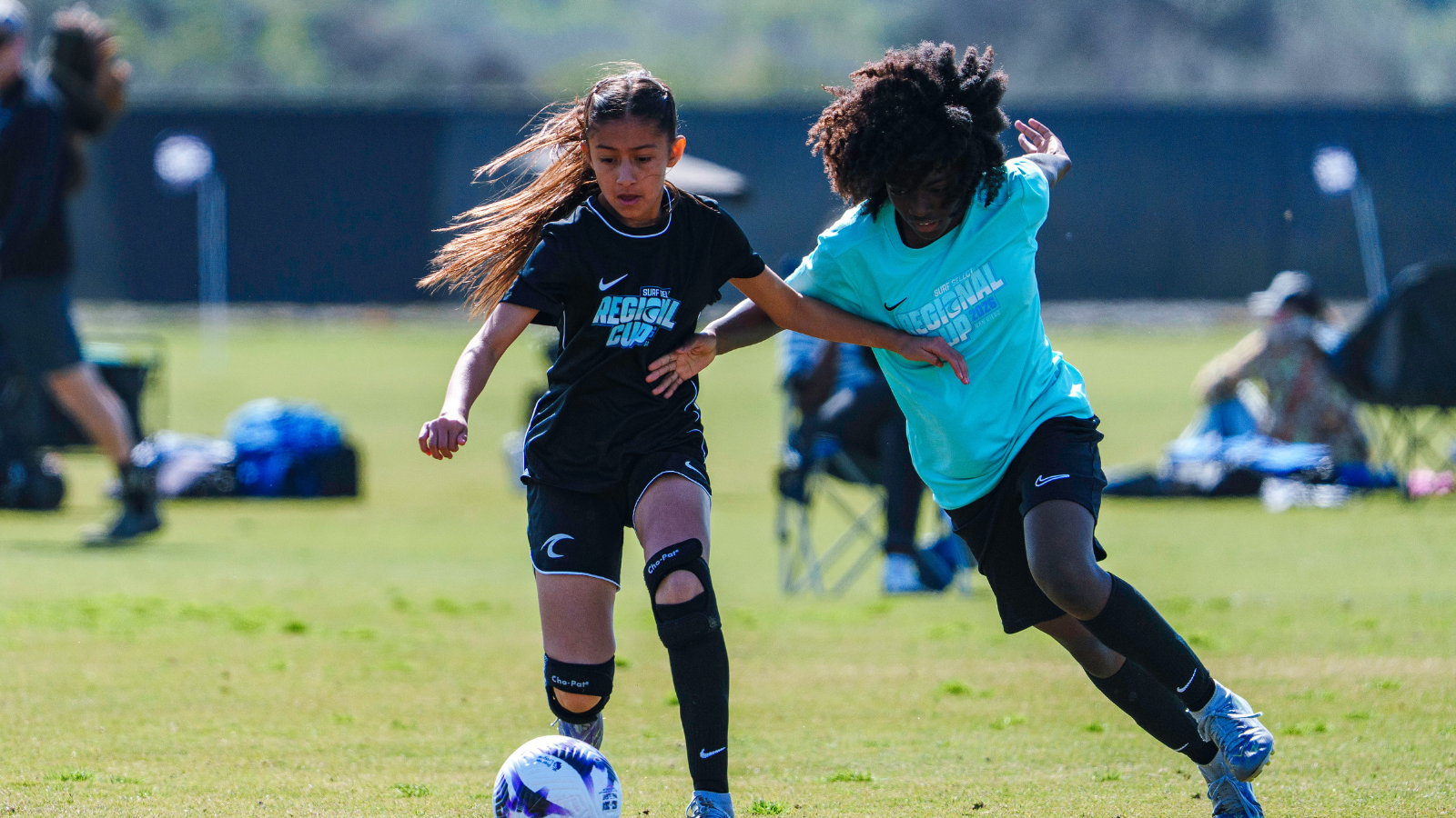 Two girls in a soccer match chase a ball on a sunny field, one in black kit and the other in turquoise, both reaching for possession.