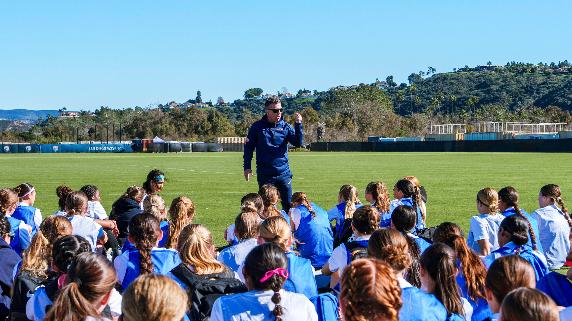 Coach addressing a group of young players seated on a sunny soccer field with hills in the background, during practice