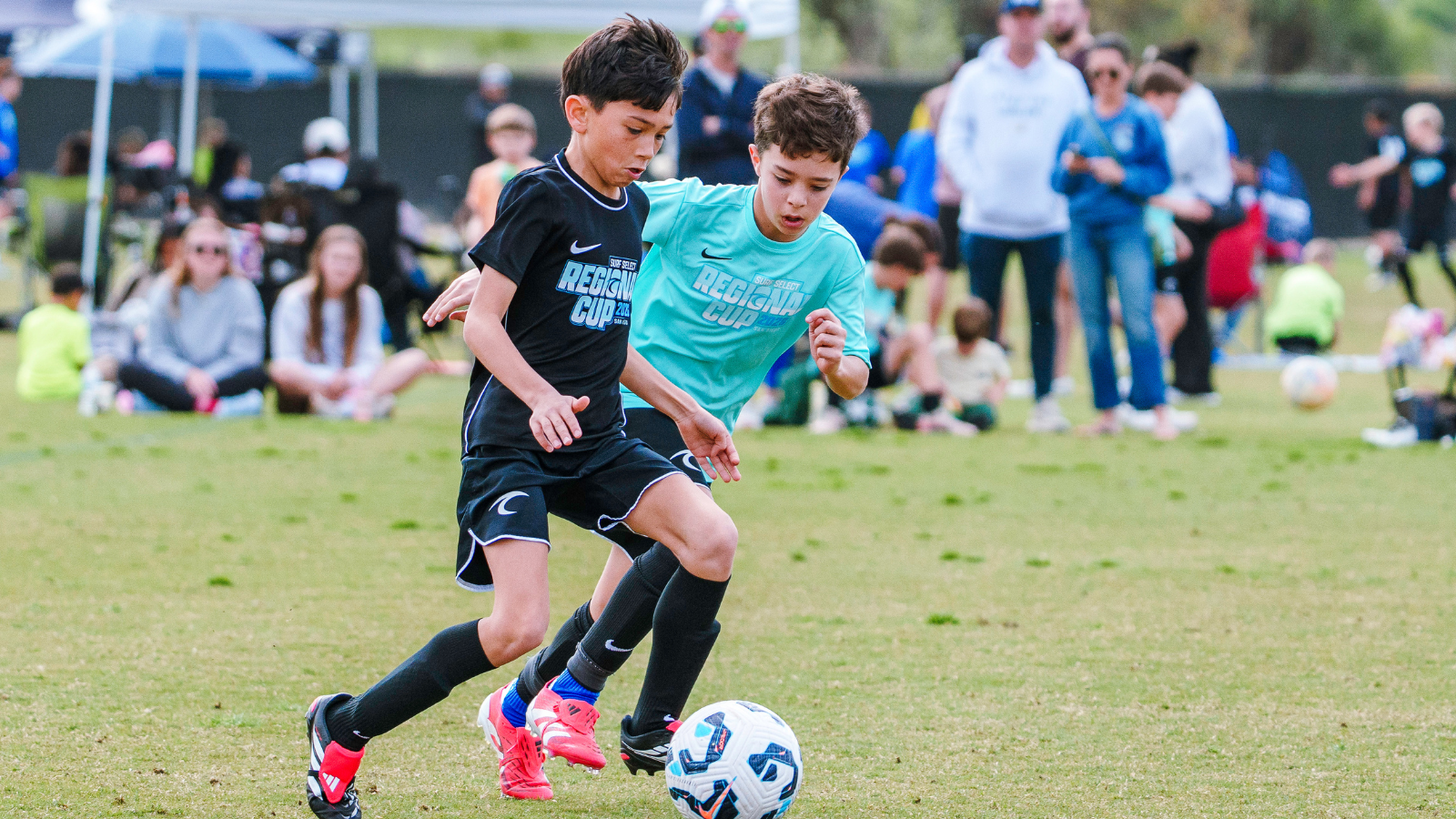 Two young boys compete for a soccer ball on a grassy field, with spectators seated and standing in the background.