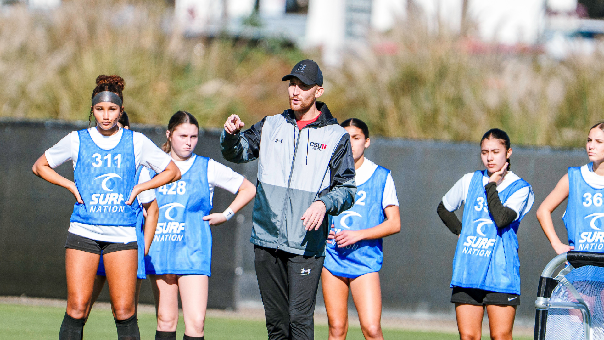 Coach giving instructions to a girls' team in blue pinnies on a sports field.