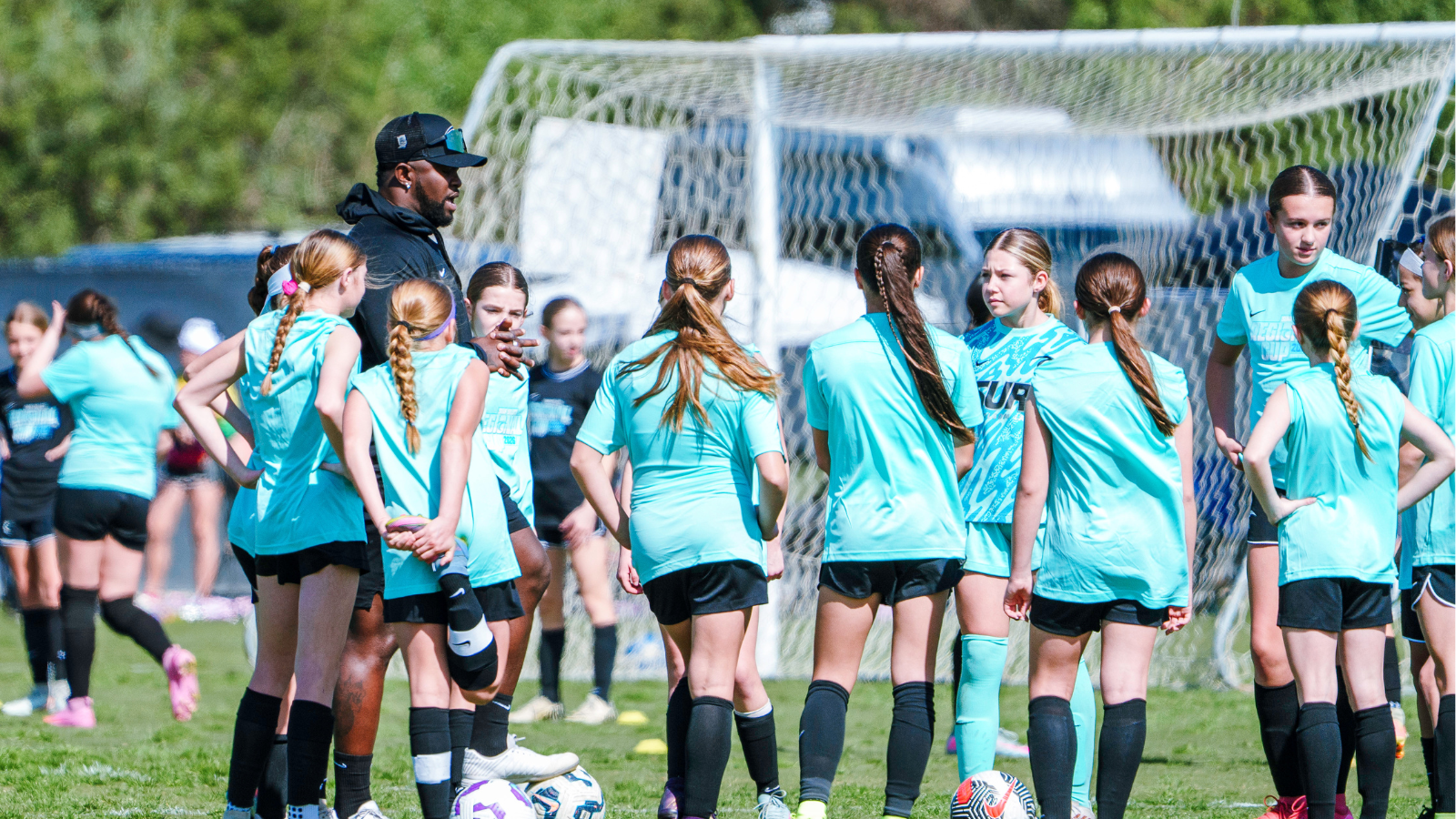 Youth girls soccer team gathers around coach during practice on a sunny field by the goal