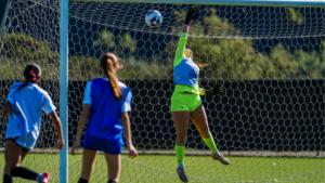 Female goalkeeper in neon green kit leaps to save a ball at the goal while two players in blue defend nearby.