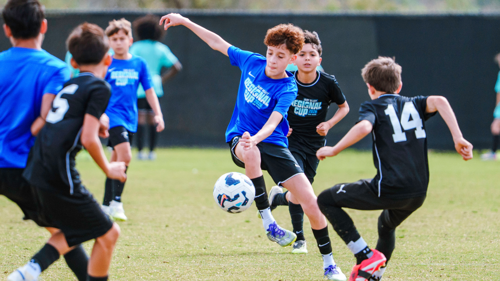 Youth soccer match with a boy in a blue shirt kicking a ball as others in blue and black jerseys chase on a grass field