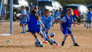 Young girls playing soccer on a dirt field near the goal, with three players chasing the ball. Two wear blue bibs, one in white with '174'.