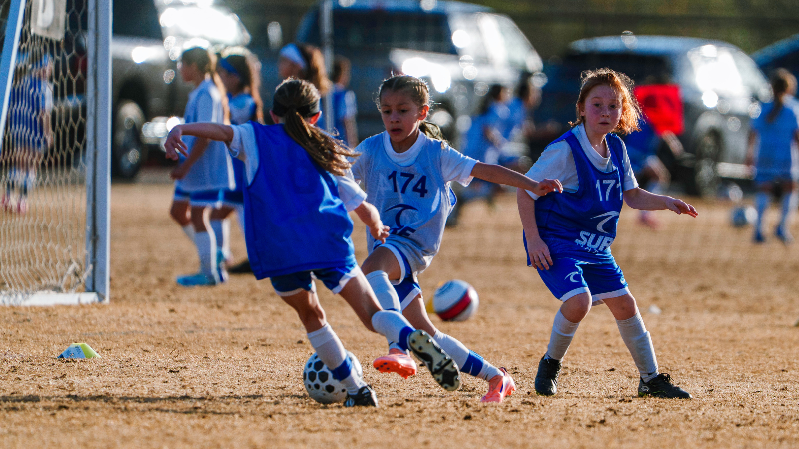 Young girls playing soccer on a dirt field near the goal, with three players chasing the ball. Two wear blue bibs, one in white with '174'.