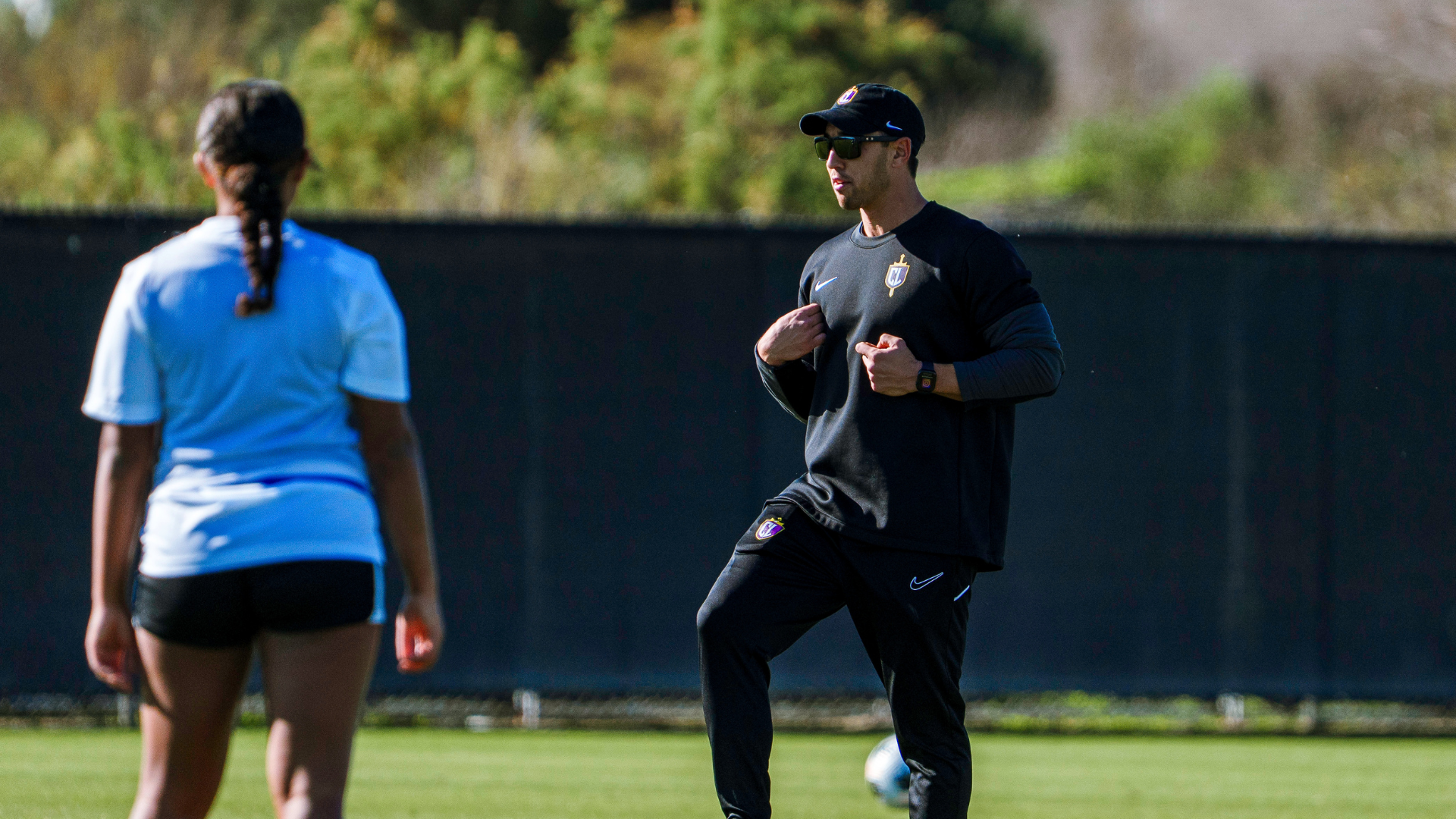 Male athlete in black training gear with cap and sunglasses demonstrates a drill on a grassy practice field, while a teammate in blue watches nearby.
