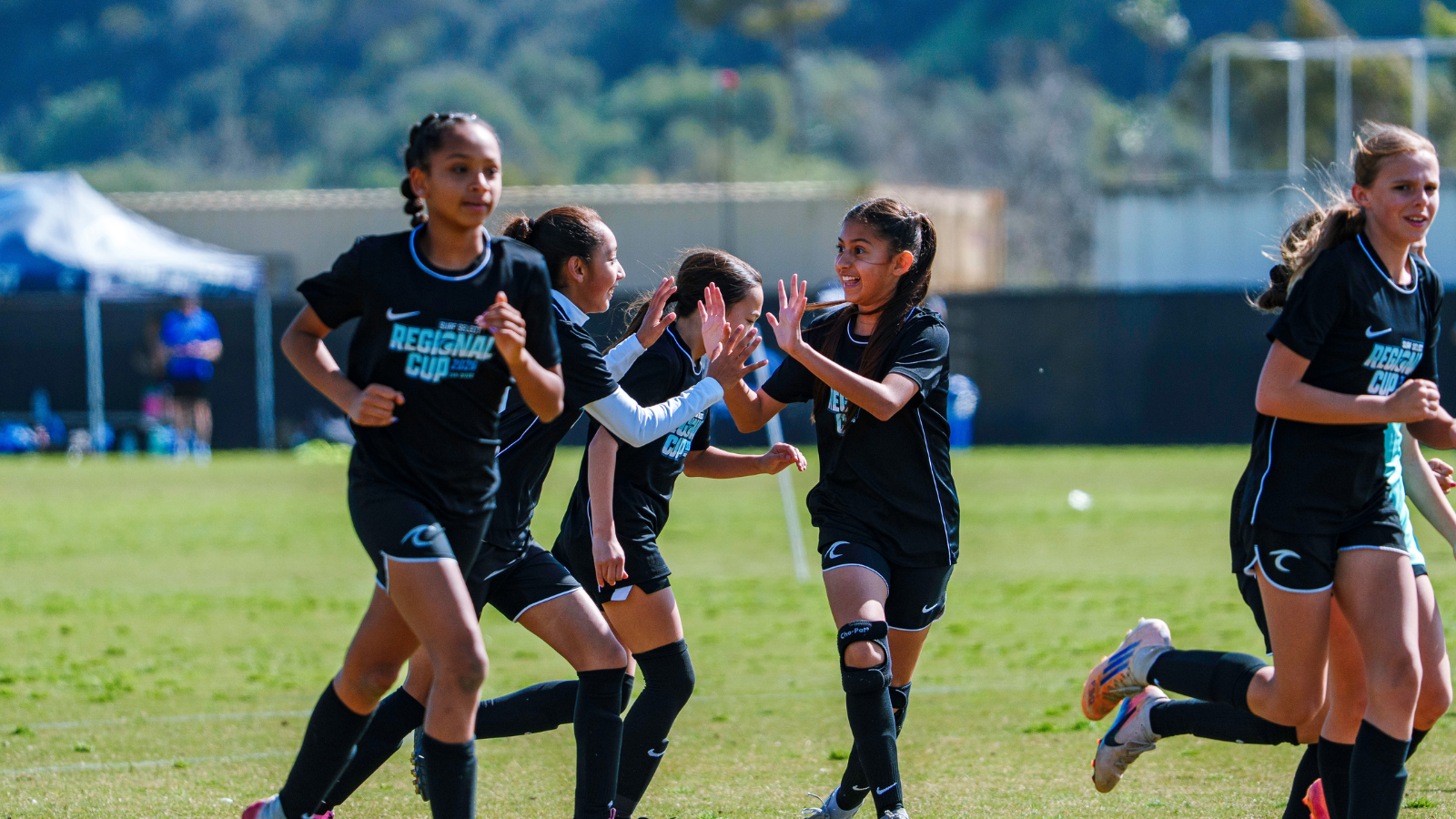 Youth girls' soccer team in black uniforms celebrate with high-fives on a sunny field.