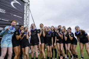 Group of young female athletes posing with medals after a game, standing on a field beside a large event banner and metal scaffolding, smiling at the camera.