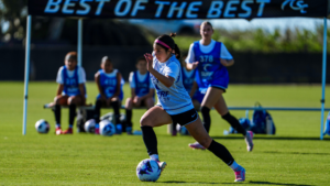 Female soccer player in a light blue jersey dribbles the ball on a sunny field, with teammates on the bench in the background.