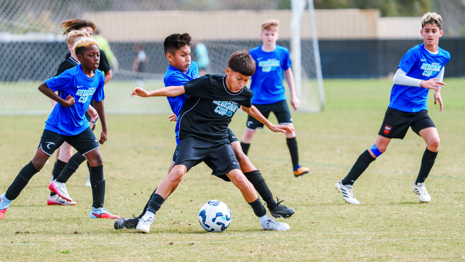 Youth soccer match: a boy in black jersey controls the ball as blue-jersey opponents reach in for a tackle on a grassy field with a goal in the background