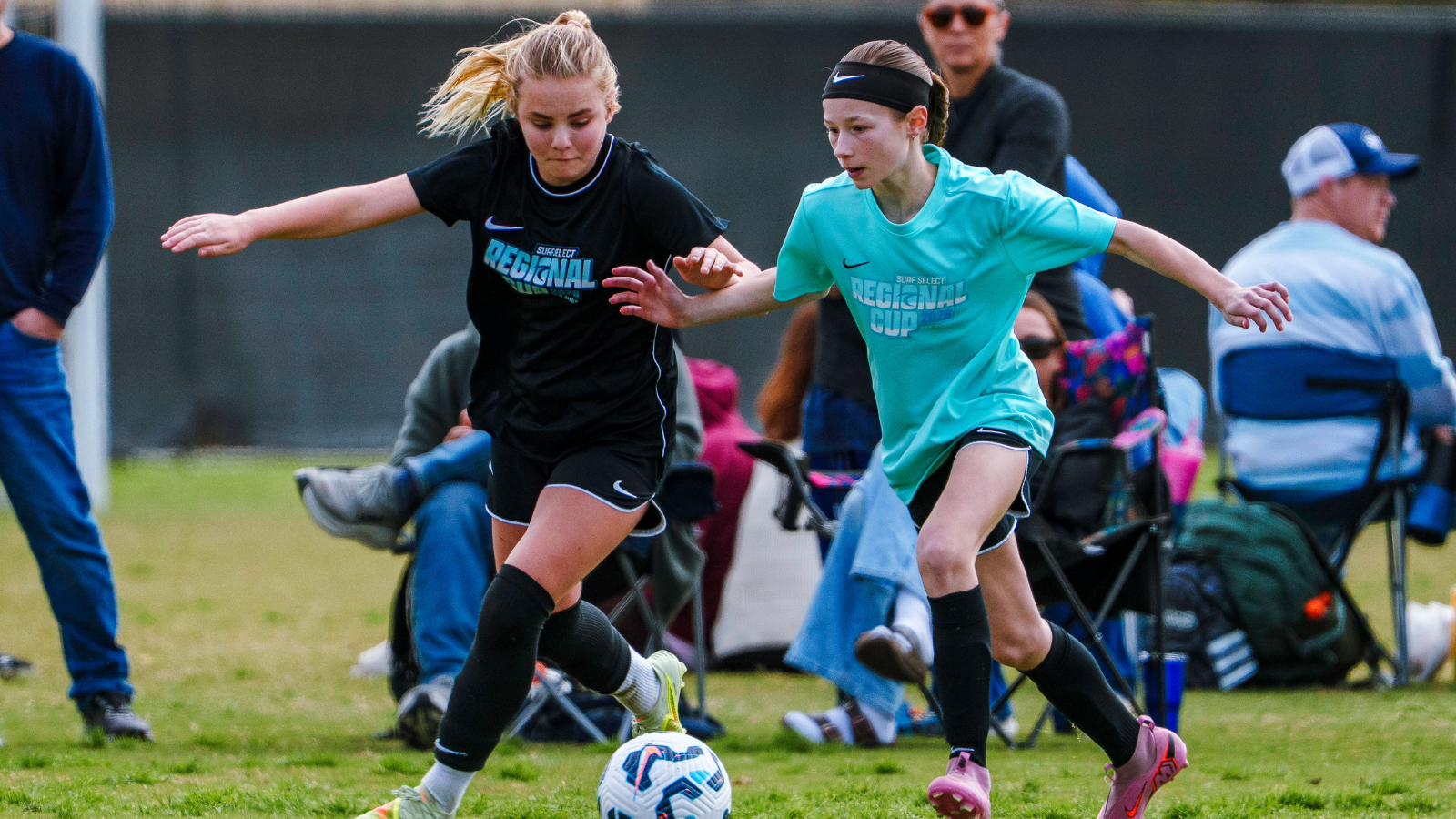 Two young girls compete for a soccer ball during a match on a grassy field, one in a black kit and the other in turquoise, both focused on the ball.