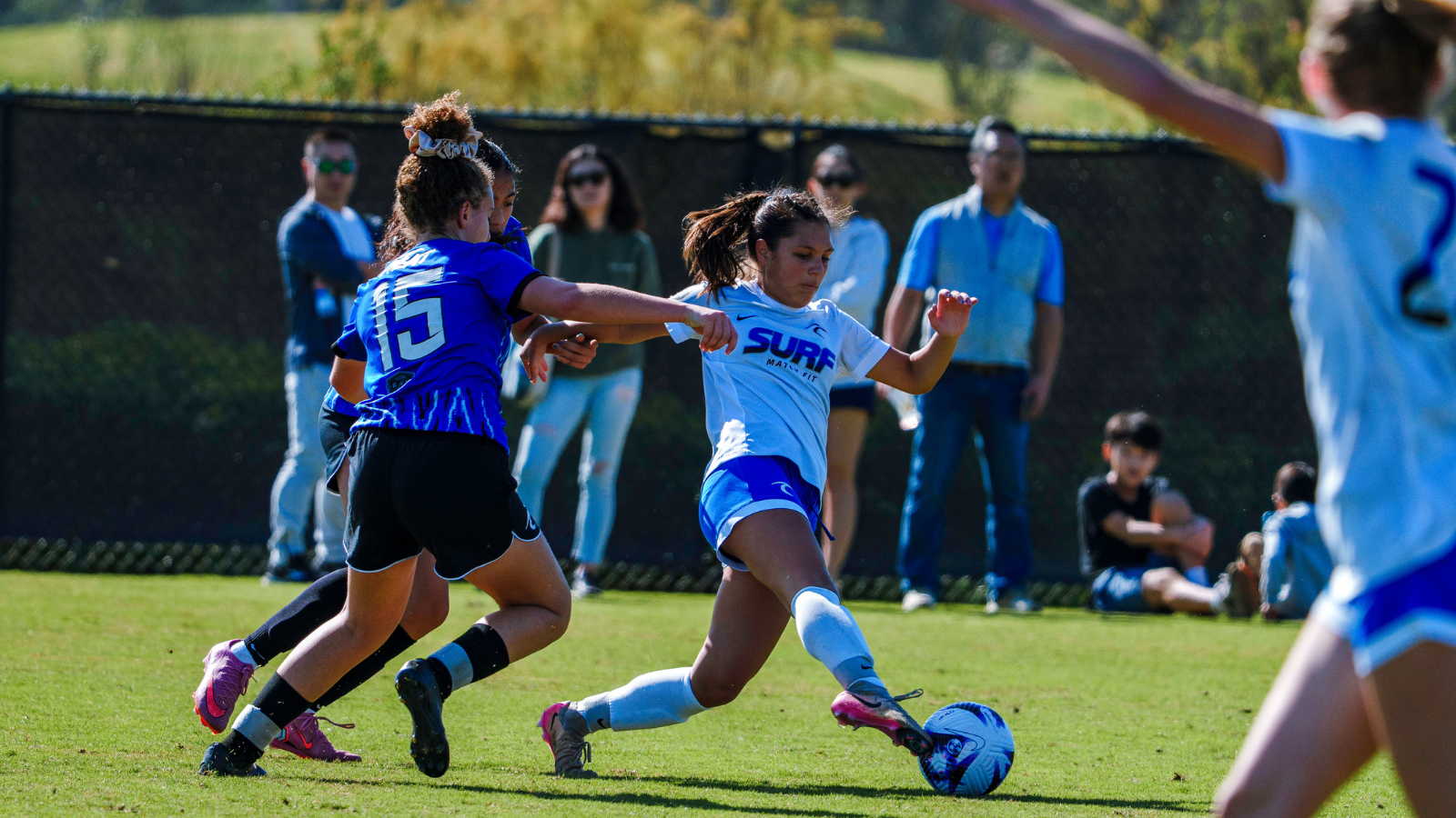 Girl soccer player in white jersey kicks a blue ball while a defender in blue tries to block on a sunny field.