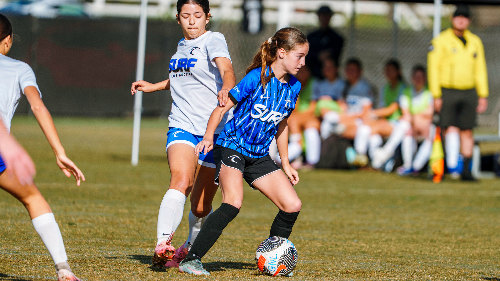Young female soccer player in blue jersey dribbles a ball while being challenged by a white-jersey opponent on a sunny field; spectators and a referee sit in the background.