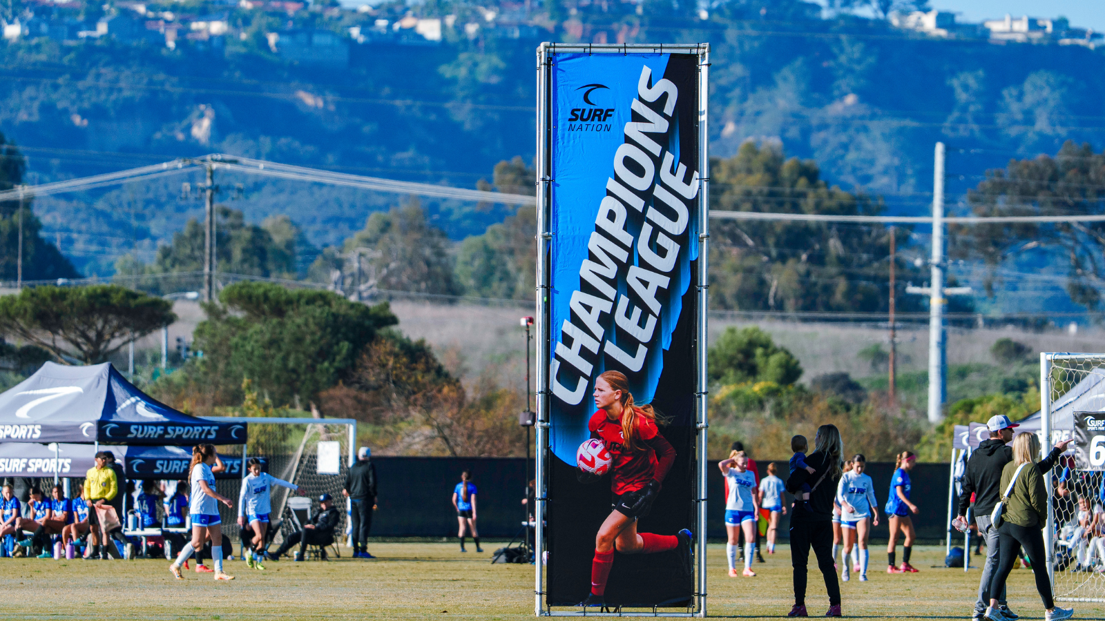 Young female soccer goalie in red runs with a ball beside a tall blue 'Champions League' banner on a sunny field. Players and tents are in the background.