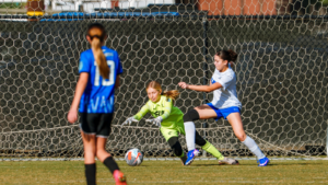 Girls' soccer near the goal as the goalkeeper in neon green dives to stop the ball while a white-team player approaches.