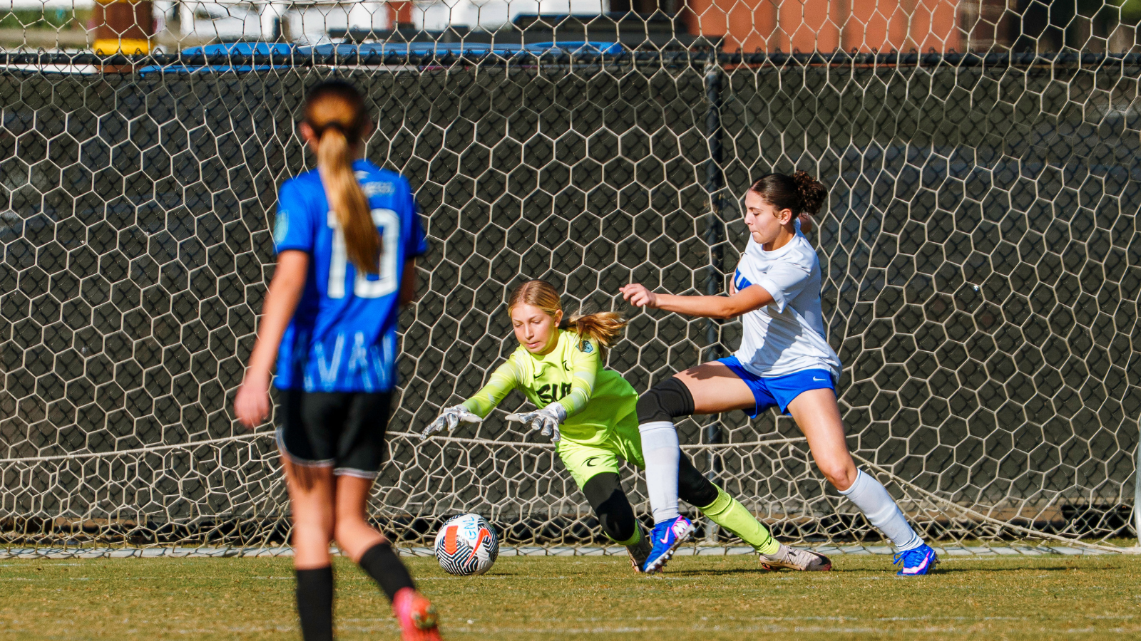 Girls' soccer near the goal as the goalkeeper in neon green dives to stop the ball while a white-team player approaches.