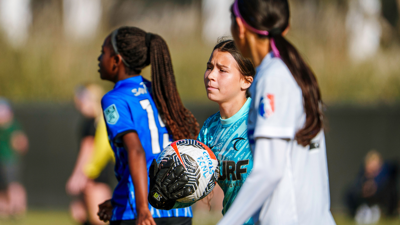 Young female soccer players on a field, with one girl in teal holding a ball and preparing to pass or throw in during a game of youth soccer.
