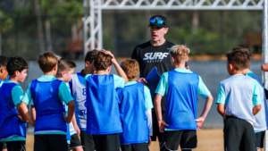 Coach giving instructions to a group of young soccer players during outdoor practice on a sunny field, clipboard in hand.