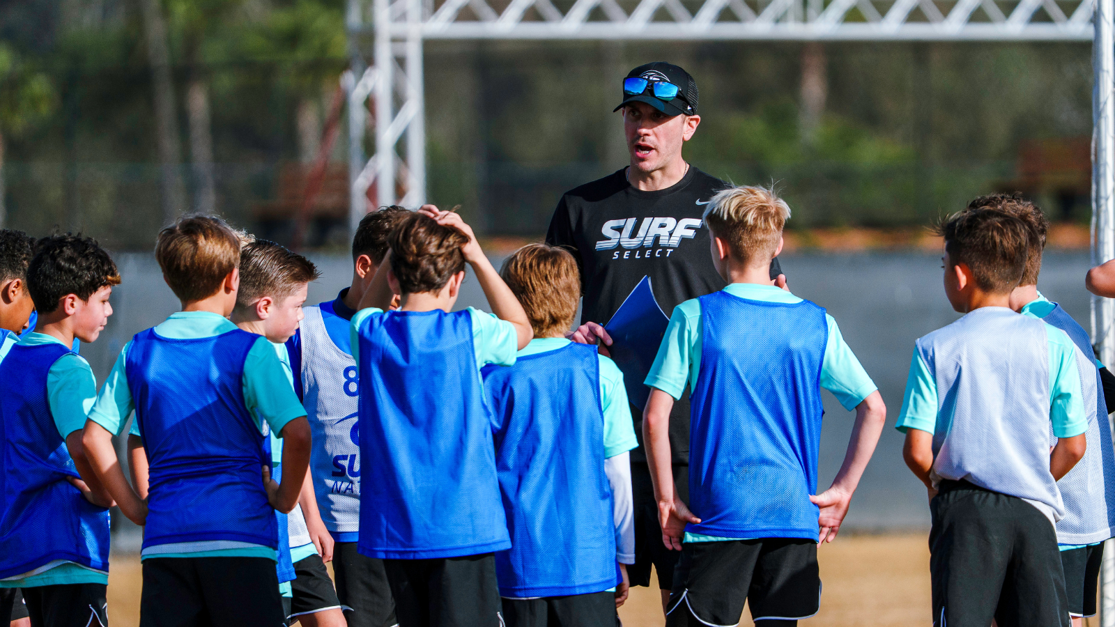 Coach giving instructions to a group of young soccer players during outdoor practice on a sunny field, clipboard in hand.