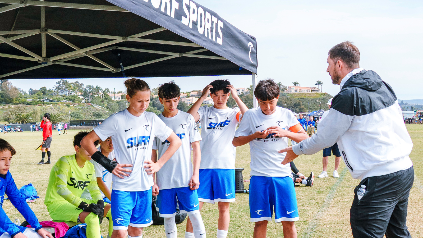 Coach giving instructions to a youth soccer team in white and blue uniforms under a canopy before a match
