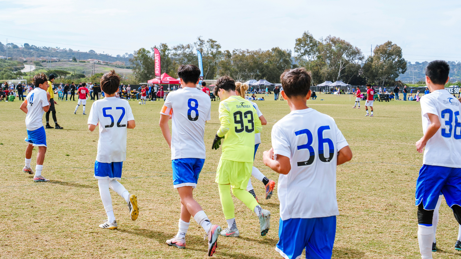 Youth soccer game on a sunny field with kids in white and blue uniforms; a neon‑green goalie stands near the action as spectators and tents line the background.
