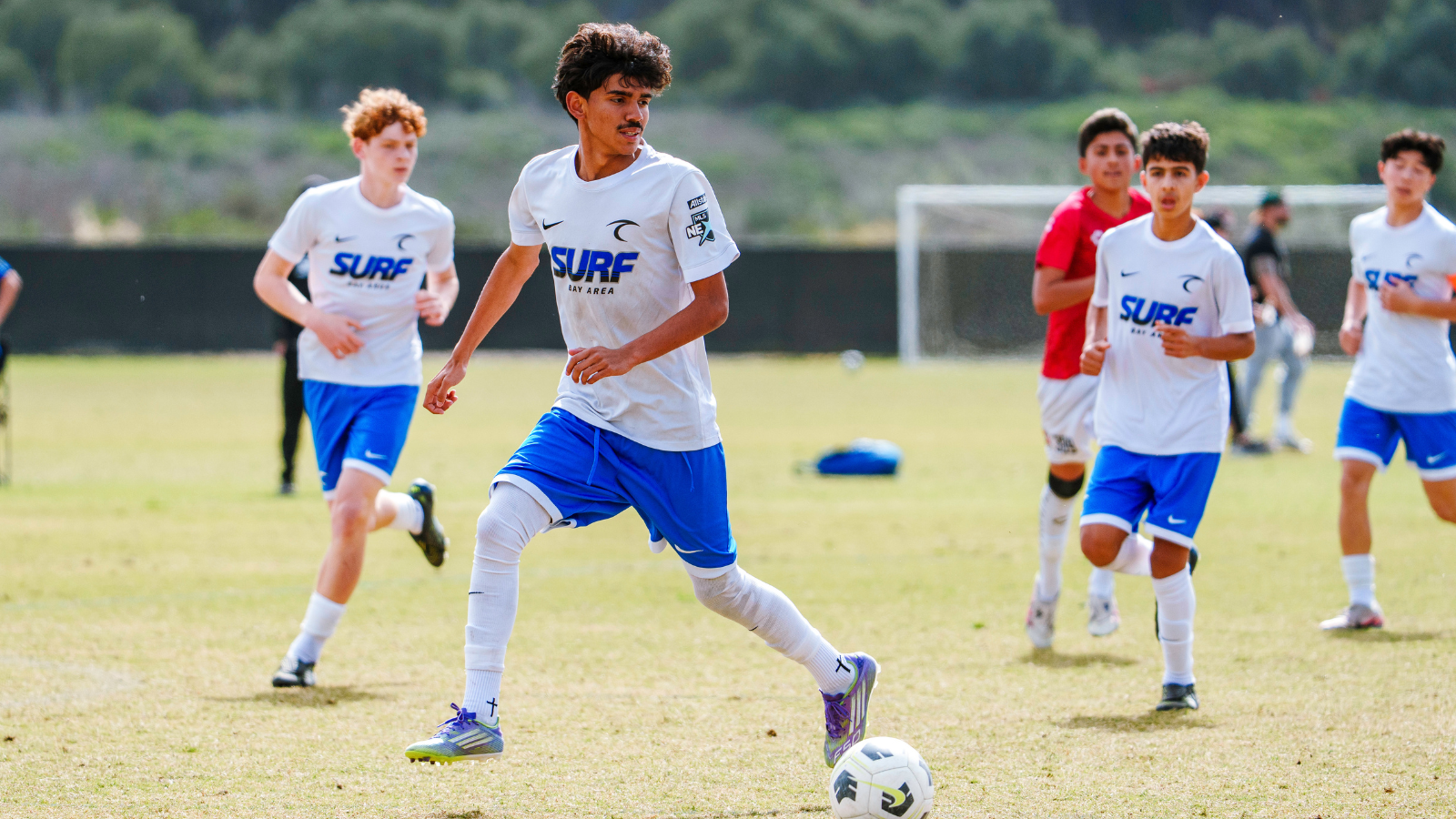 Young soccer players in white jerseys and blue shorts run on a sunny field; central player dribbles the ball toward the goal.