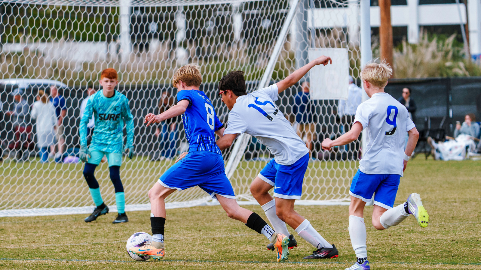 Youth soccer game with a blue team player controlling the ball near the goal as white-team players close in; goalkeeper in turquoise stands ready in the net behind them.