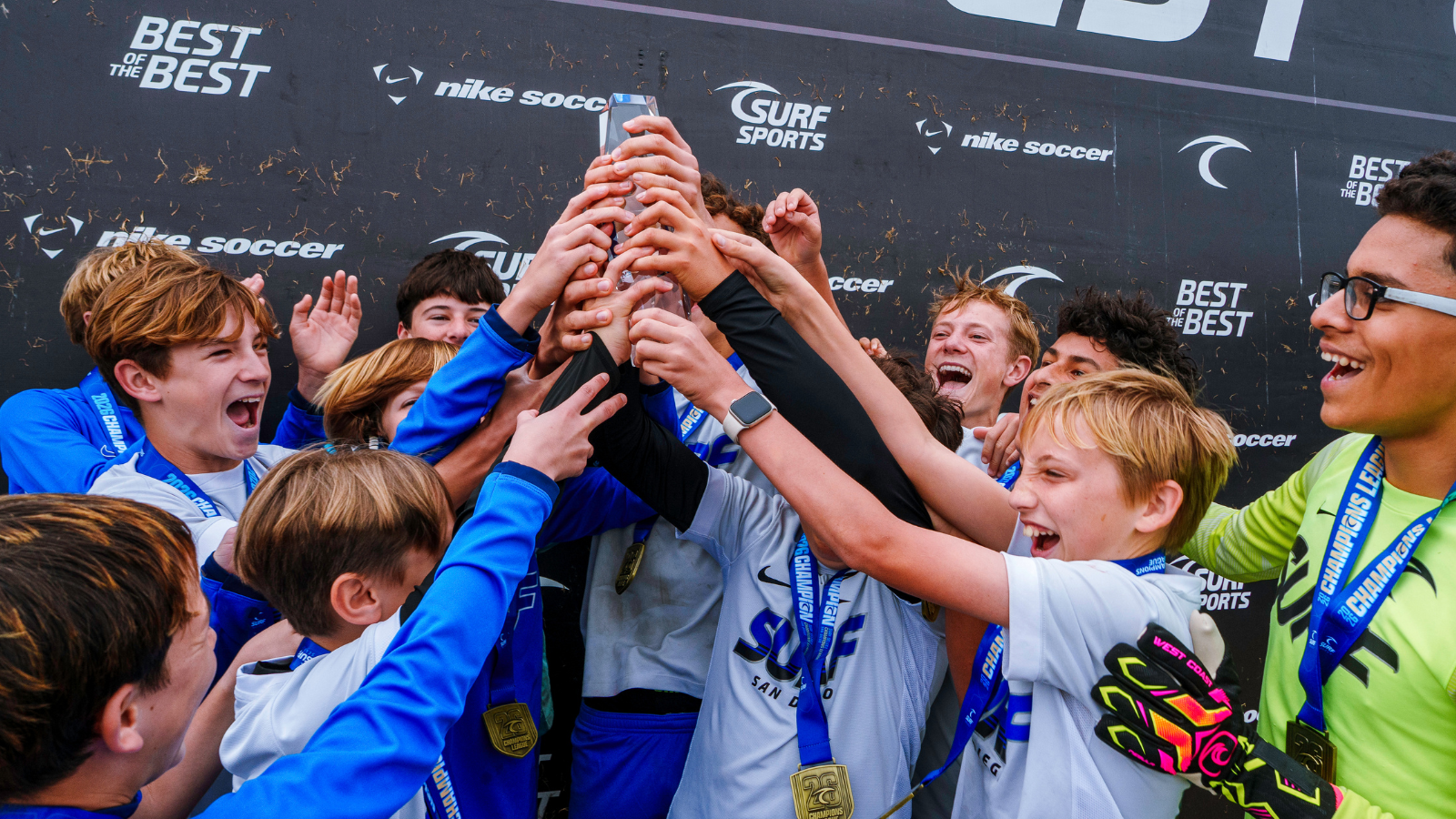 Youth soccer team celebrates with a trophy aloft, medals around their necks on a dark backdrop.
