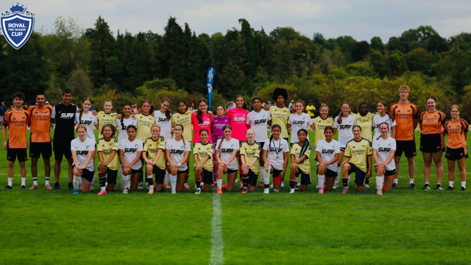 Group of young female soccer players posing for a team photo on a grassy field, wearing white, yellow, and orange jerseys with a Royal Pre Season Cup crest in the top-left corner.