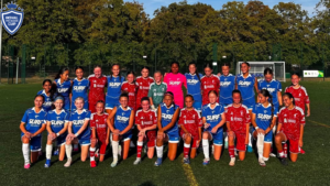 Group photo of a girls' soccer team on a field, wearing blue and red uniforms, with a crest logo in the top-left corner.