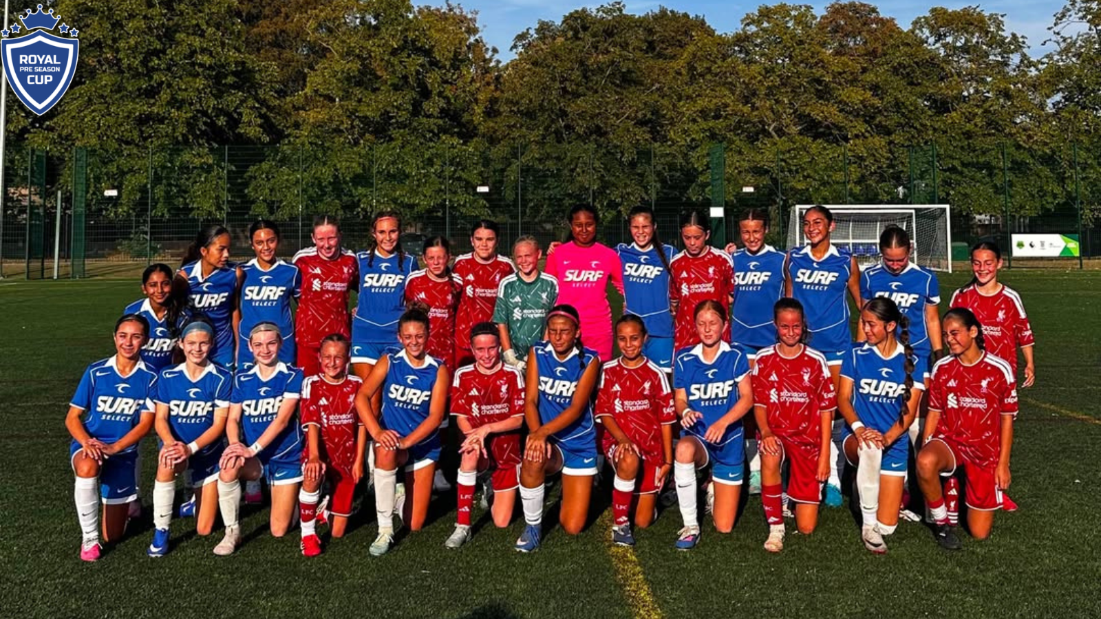 Group photo of a girls' soccer team on a field, wearing blue and red uniforms, with a crest logo in the top-left corner.