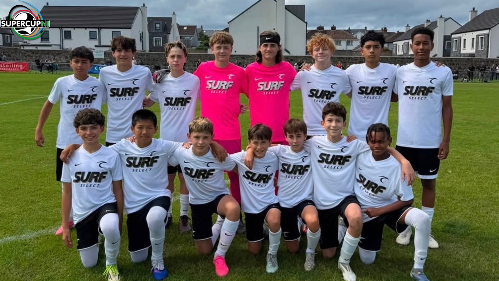 Youth soccer team posing for a group photo on a grassy field, players in white jerseys with 'SURF SELECT' and two pink-jersey goalkeepers in the middle, backdrop of houses and a small stadium.
