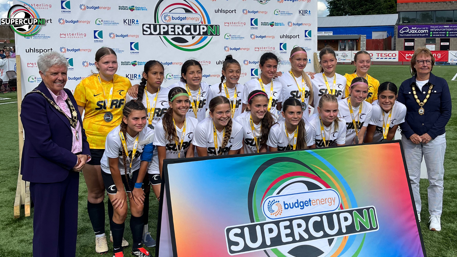 Girls soccer team in white jerseys with medals pose for a group photo in front of a bright SUPER CUP NI backdrop, flanked by two officials on each side.