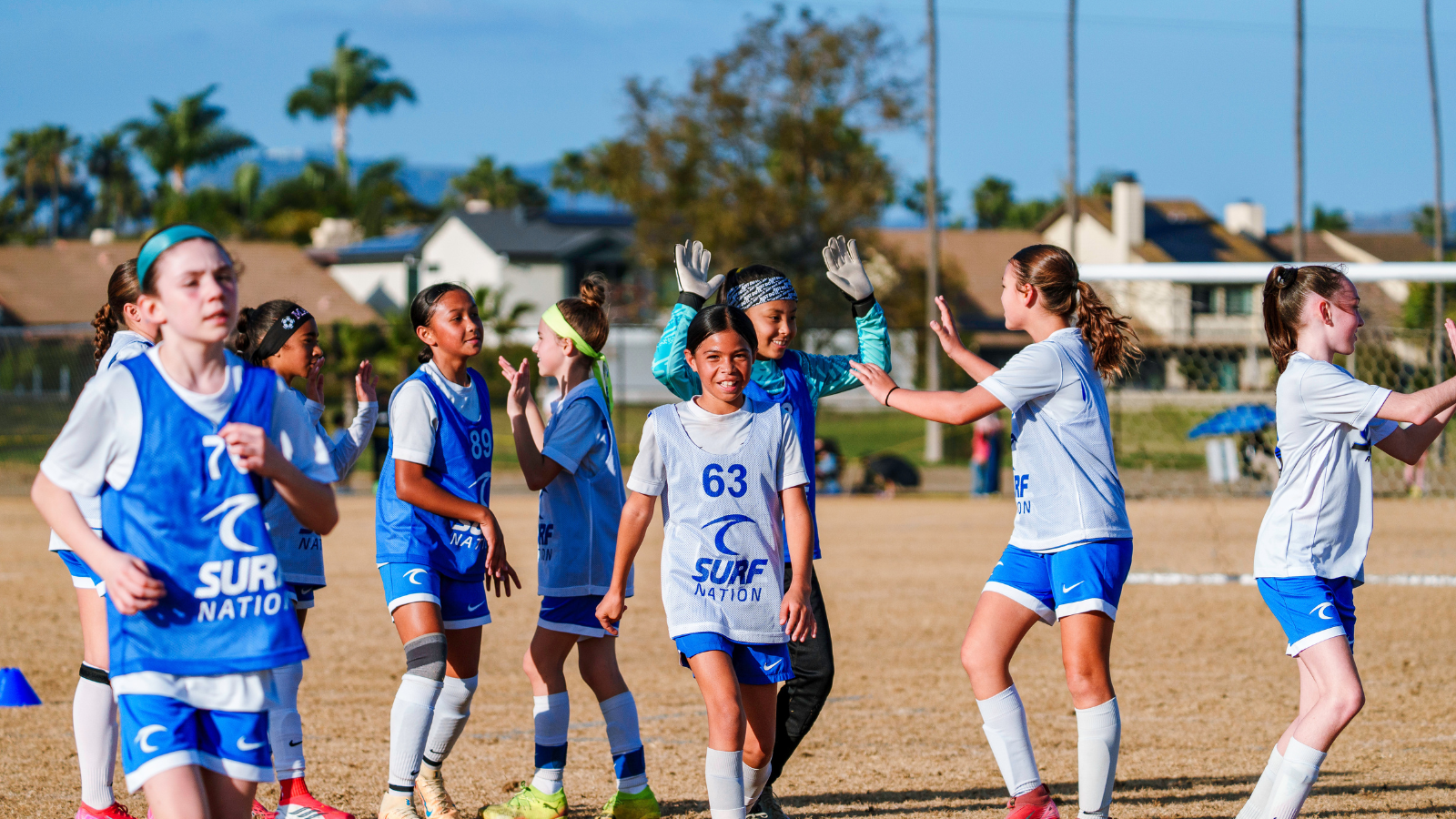 Young girls in blue and white jerseys celebrate with high-fives on a sunny sports field; central player wearing #63 smiles at the camera.