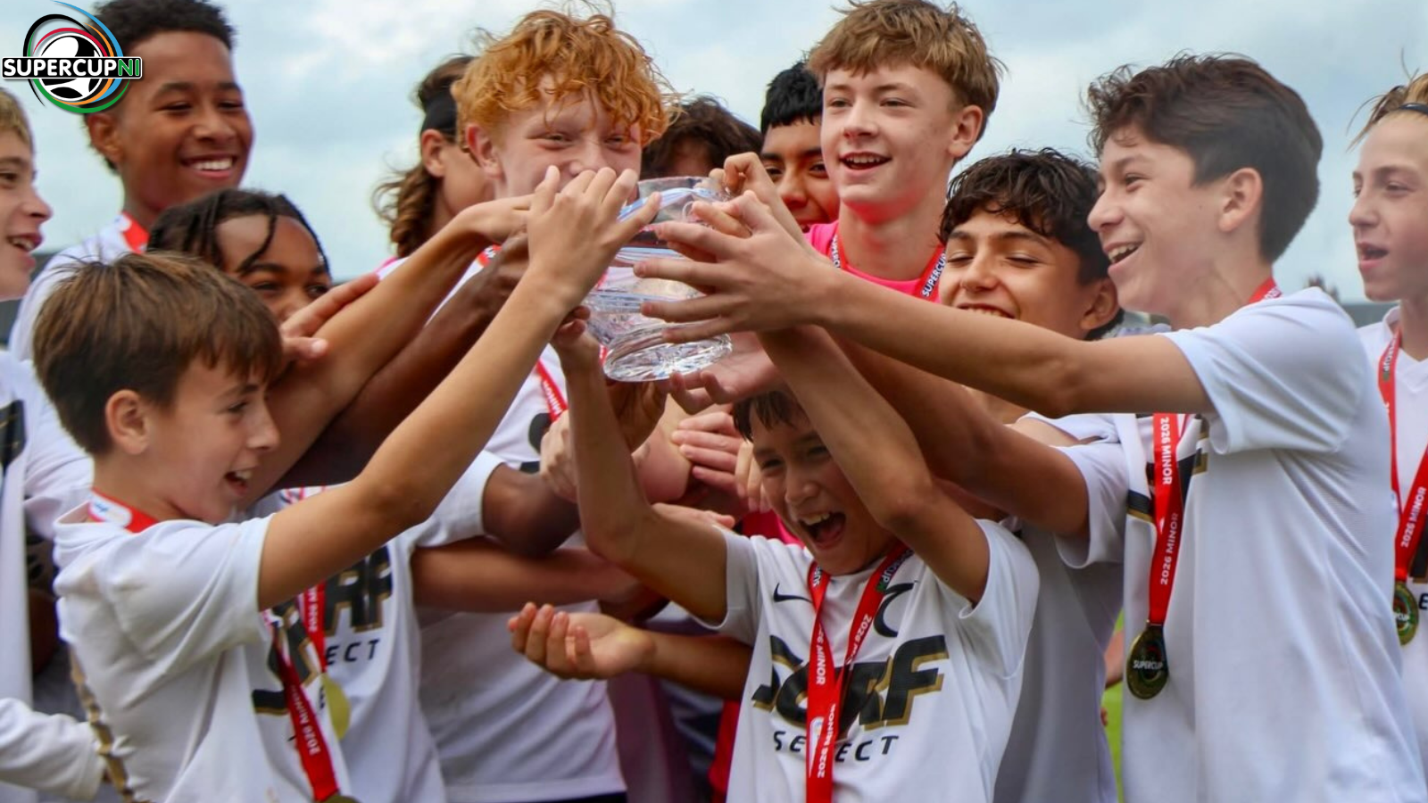 Young soccer players in white jerseys celebrate, lifting a trophy together with medals around their necks; SuperCup NI logo visible in corner.