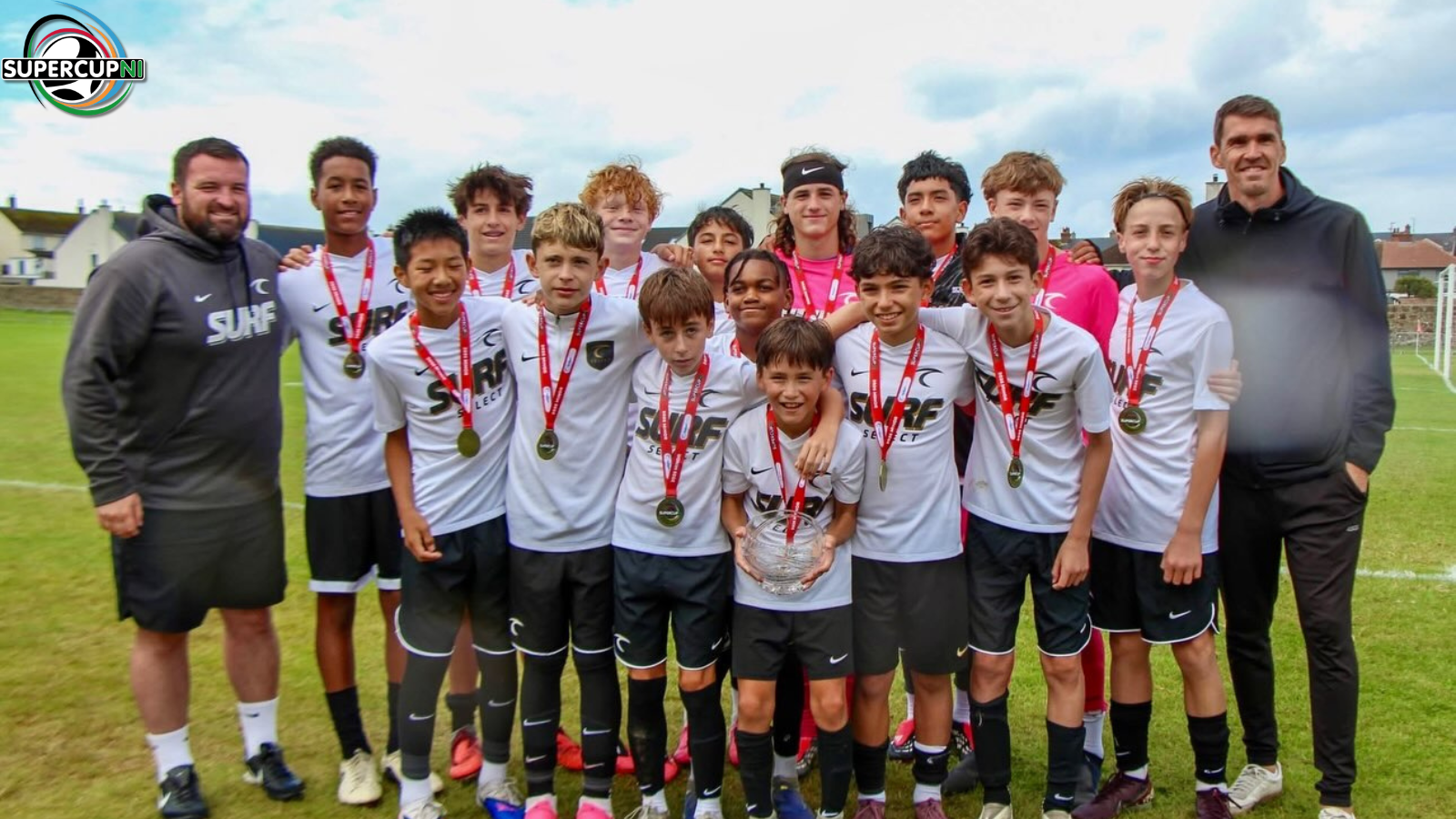 Youth soccer team pose on field with medals after victory, holding a trophy, flanked by coaches and a logo in the corner (SuperCupNI).