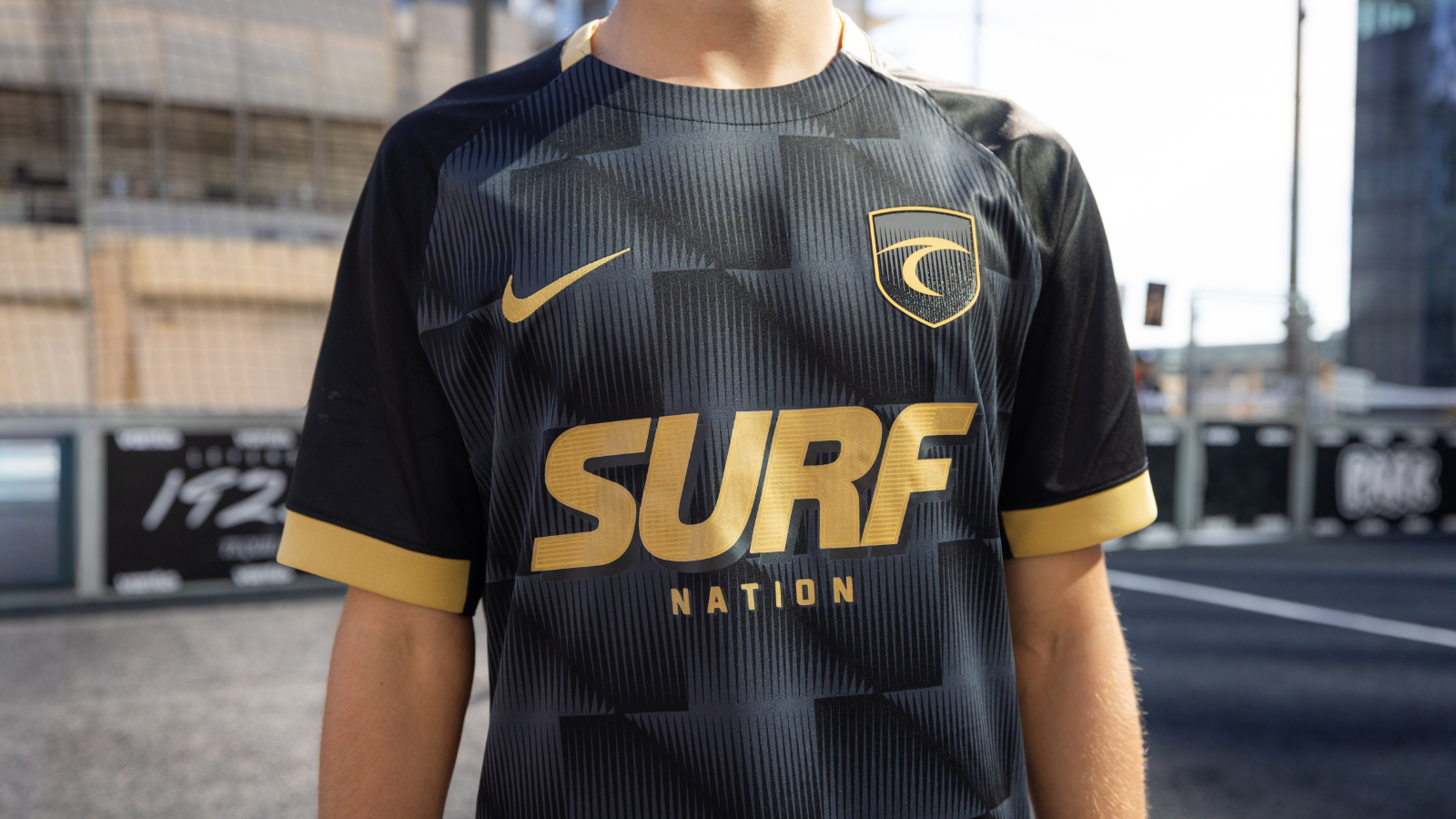 Person wearing a black and gold sports jersey with a large 'SURE NATION' logo and crest on the chest, outdoors in an urban setting.