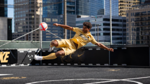 Male futsal player in a yellow kit performing a bicycle kick on a rooftop urban court with a red-and-white ball nearby.