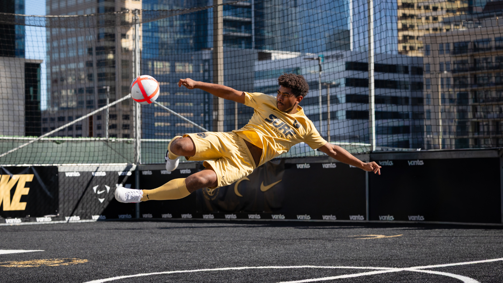 Male futsal player in a yellow kit performing a bicycle kick on a rooftop urban court with a red-and-white ball nearby.
