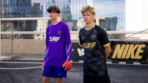 Two teenage soccer players stand on an urban rooftop futsal court in purple and black SURF kits, with a goal and net behind them. Gloves are orange on the player in purple.